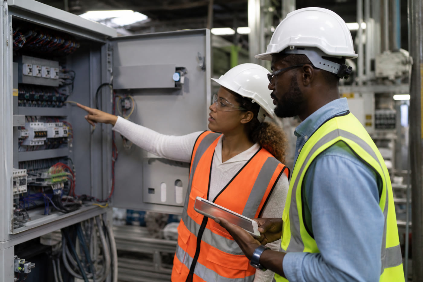 A woman and a man in hard hats and safety vests reviewing an open electrical panel, one holding a tablet.